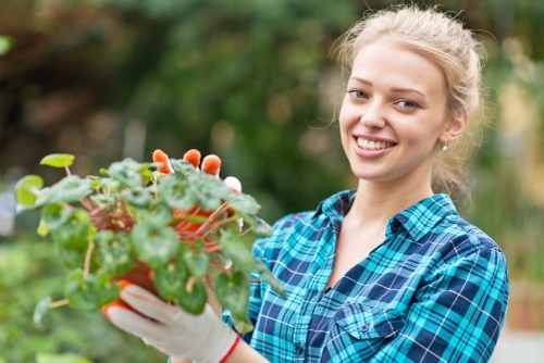 A young woman with blonde hair tied back, smiling while holding a potted plant with green and reddish leaves, standing outdoors in a garden environment. The background features a variety of green foliage, including trees and shrubs, with natural sunlight illuminating the scene. She is wearing a blue and black checkered shirt and gardening gloves, suggesting she is engaged in gardening activities. The garden setting includes a well-maintained lawn area and various plantings, typical of outdoor spaces in Hammersmith. This image exemplifies outdoor gardening work and outdoor maintenance that a landscaping service such as Landscapers Hammersmith might undertake, especially in residential gardens in London, including patios, lawns, and plant care.