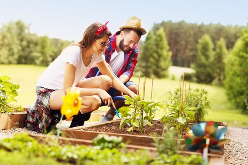 The image depicts a young woman and a man working together in a garden, tending to raised flower beds with small green plants and budding flowers. The garden features a neatly maintained grassy lawn in the background, bordered by low brick walls and supported by wooden edging around the planting areas. The scene is set outdoors on a bright, sunny day with clear skies, surrounded by lush green trees and a natural landscape typical of suburban gardens in Hammersmith. The garden includes soil beds with dark, rich earth, fresh green foliage, and emerging blooms, indicating ongoing planting and cultivation. The individuals are casually dressed for gardening, with the woman wearing a white top, plaid shorts, and a pink headband, and the man in a plaid shirt and straw hat, both engaged in nurturing young plants. This scene reflects typical gardening activities that enhance outdoor spaces, aligning with services offered by companies like Landscapers Hammersmith, focusing on garden planting, maintenance, and landscape improvements in the local area.
