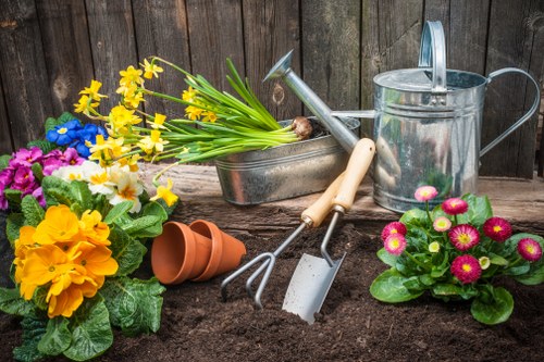 An outdoor garden scene in a neatly maintained backyard with a wooden fence in the background. The foreground features a freshly dug garden bed with dark, rich soil, ready for planting. To the left, there are vibrant flowering plants including yellow primroses, blue and pink pansies, and orange primulas, all with healthy green foliage. A small terracotta plant pot lies on its side near the flowers. In the centre, garden tools such as a hand rake, trowel, and garden fork, along with a watering can, are arranged on the soil, indicating recent planting or soil preparation work. The garden area appears well-organized and ready for further gardening activities, with natural light highlighting the bright colors of the flowers and the textures of the tools and soil. The scene reflects a typical, cultivated outdoor space suitable for gardening services offered by local landscapers, making it relevant to services in Hammersmith or surrounding areas.