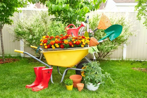 A colorful garden scene featuring a yellow wheelbarrow filled with vibrant red and yellow flowering plants positioned on a well-maintained grassy lawn. Surrounding the wheelbarrow are gardening tools and accessories, including a red watering can, green and orange gardening spades, a small white planter, and several empty terracotta pots arranged on the grass. In the background, there are lush green hedges, leafy trees, and a white wooden fence that encloses the outdoor space, which appears to be part of a residential garden in Hammersmith. The scene suggests an active gardening area prepared for planting and outdoor maintenance, with natural daylight indicating a clear, pleasant day suitable for gardening services. This setup aligns with the typical landscaping and garden care tasks offered by Landscapers Hammersmith, emphasizing well-kept lawns, plant cultivation, and garden organisation across the local area.