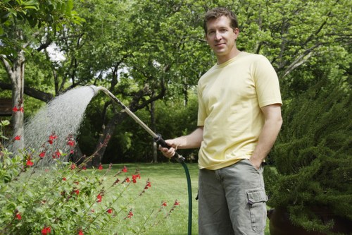 A man standing in a lush, well-maintained garden in Hammersmith, England, holding a garden hose and watering a flower bed with red blooms. The garden features a neatly trimmed lawn, a variety of shrubs, and mature trees providing shade. In the background, a wooden hedge and a paved patio area are visible, with natural sunlight illuminating the scene on a clear day. The environment reflects typical outdoor garden care in the local area, with vibrant greenery and colorful flowers. Landscaping services by Landscapers Hammersmith likely include plant and lawn maintenance, garden watering, and outdoor cleaning to keep such gardens thriving.