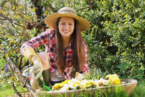 A young woman with long brown hair wearing a straw sunhat, plaid shirt, and gardening gloves is kneeling in a lush garden. She is tending to a large, round, wooden planter filled with bright yellow and white flowering plants. Behind her, various green plants, shrubs, and a tree with twisted branches provide a natural backdrop. The garden features a mix of dense foliage and flowering plants, with some soil visible near the base of the plants. The scene appears to be outdoors on a sunny day, with natural lighting highlighting the vibrant greens and floral colours, creating a serene and productive gardening environment suited to sustainable outdoor care by professionals like Landscapers Hammersmith in the Hammersmith area, close to postcode W6, supporting local gardening and landscaping services.
