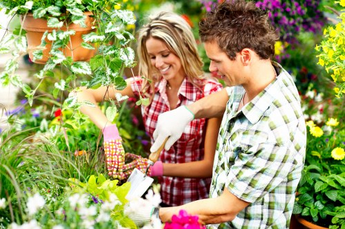 In a vibrant garden environment, a young woman and man are engaged in gardening activities amidst lush greenery and colourful flowering plants. The woman, wearing pink gardening gloves and a red checkered shirt, is smiling as she holds a small gardening tool and tends to a leafy plant. The man, dressed in a white and green checked shirt and white gloves, is focused on the planting or pruning task, standing close to her. The garden features various textured elements, including dense green grass, a neatly maintained flower bed with pink, purple, and yellow blossoms, and potted plants hanging from above. The background displays taller shrubs and flowering bushes, adding depth to the landscaped yard. The scene suggests a well-maintained outdoor space typical of residential gardens in Hammersmith, with natural daylight illuminating the scene, highlighting the vibrant colours and textures of the plants. This image exemplifies professional gardening activities by Landscapers Hammersmith, showcasing the care and attention involved in maintaining attractive outdoor spaces in London suburbs, suitable for enhancing garden aesthetics and outdoor enjoyment.