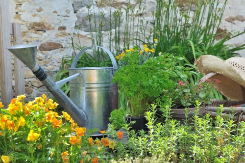 The image depicts a small garden area with a variety of plants and gardening tools set against a rustic stone wall. In the foreground, there are bright yellow flowering plants adjacent to a patch of green grass with dense, evenly cut turf. Behind the flowers, a weathered metal watering can with a curved handle and spout is placed among potted and planted greenery, including a bushy plant with light green, serrated leaves. To the right, there is a section of soil bordered by small plants and possibly a decorative border, with a straw hat resting on the edge of a garden feature. Tall, slender green grass or ornamental plants grow vertically in the background, adding height and texture to the scene. The overall environment appears to be an outdoor garden or landscaped yard in a residential area in Hammersmith, suitable for gardening and landscaping services that maintain and enhance outdoor spaces. The scene suggests a well-kept, cheerful garden ready for planting or ongoing maintenance, with natural lighting indicative of a clear, sunny day.