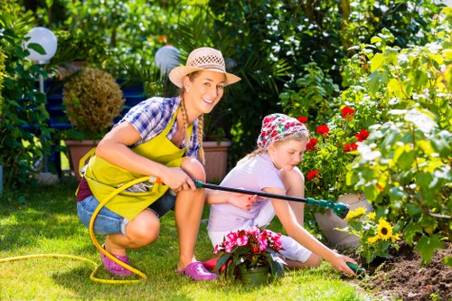 A woman and a young girl are gardening together in a lush backyard garden in Hammersmith. The woman, wearing a straw hat, blue striped shirt, and yellow apron, is kneeling on the well-maintained grass, smiling as she holds a garden hose. The girl, dressed in a pink shirt, a colorful hat, and pink shoes, is sitting on the ground, carefully planting or tending to flowering plants within a flower bed bordered by soil. The flower bed contains a variety of blooming flowers in shades of pink, red, and yellow. In the background, there are dense green shrubs, hedges, and trees, creating a private outdoor space with natural shade. Garden lanterns are visible hanging among the foliage, contributing to a welcoming atmosphere. The scene appears to be on a sunny day with bright, natural lighting, highlighting the vibrant colours and textures of the garden plants and blossoms, reflecting the outdoor gardening services provided by Landscapers Hammersmith for sustainable and appealing outdoor spaces.