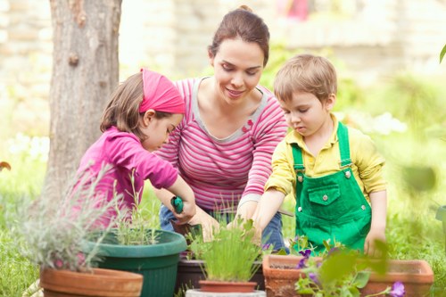 A woman with dark hair tied back, wearing a pink and white striped top, is engaged in gardening alongside a young girl dressed in a pink hoodie and a young boy in a yellow shirt with green overalls, in a lush outdoor garden. The garden features a well-maintained lawn with dense, green grass, flower beds with colorful blossoms, and potted plants arranged on a wooden surface. Behind them, there are trees with vibrant foliage and a garden fence, indicating a landscaped backyard setting. The woman and children are actively tending to various plants in terracotta and plastic pots, with tools or gardening aids potentially in use, suggesting a family gardening activity. The scene is illuminated by natural light, highlighting the lush greenery and lively colors, reflecting a positive outdoor environment typical of a residential garden in Hammersmith or the surrounding area. This image exemplifies outdoor gardening and planting activities supported by professional landscaping services, such as those offered by Landscapers Hammersmith.